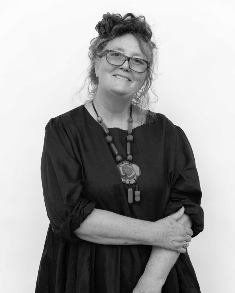 Black and white image of Beatrice Lacey, Art Consultant, with a relaxed, friendly smile. She is wearing a large stone necklace, glasses, and has her hair styled up, exuding a warm and approachable presence.